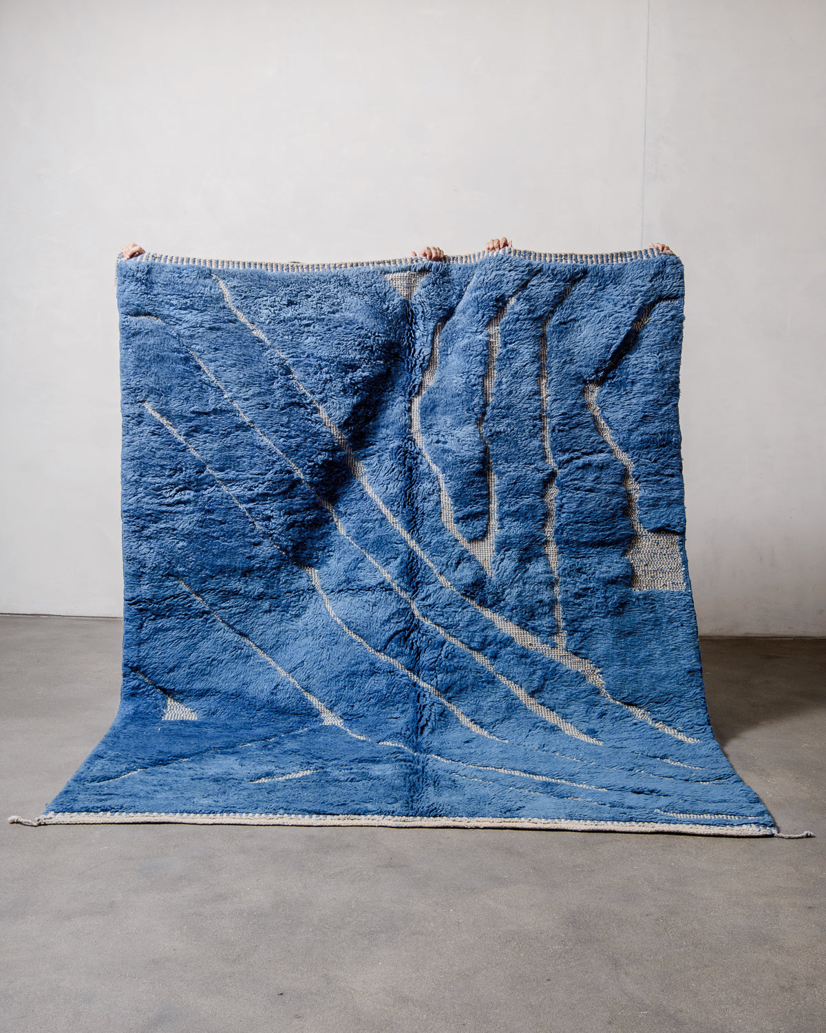 Berberlin Beni M'rirt Rug made of heavy wool pile in deep blue and cream being held up in front of a white wall and concrete floor. 