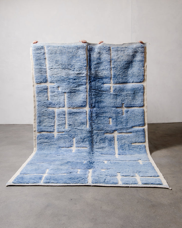 Berberlin Beni M'rirt Rug made of heavy wool pile in speckled denim blue and cream being held up in front of a white wall and concrete floor. 