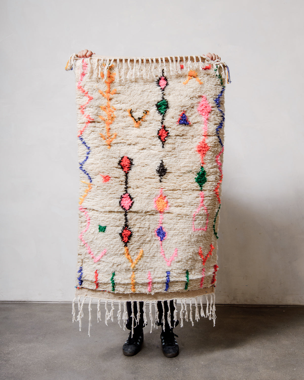 Berberlin Azilal rug in cream wool with colourful cotton  being held up in front of a white wall.