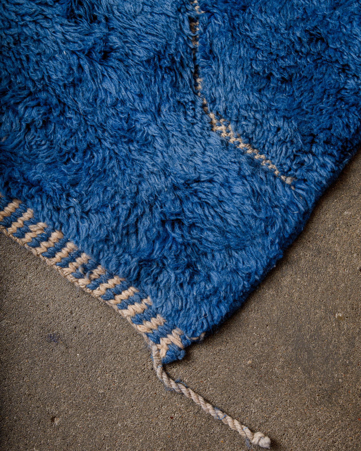 Corner of rug made of thick wool pile in deep blue and finished with tightly woven end with alternating lines of blue and cream, and a single wool tassel on the corner. 