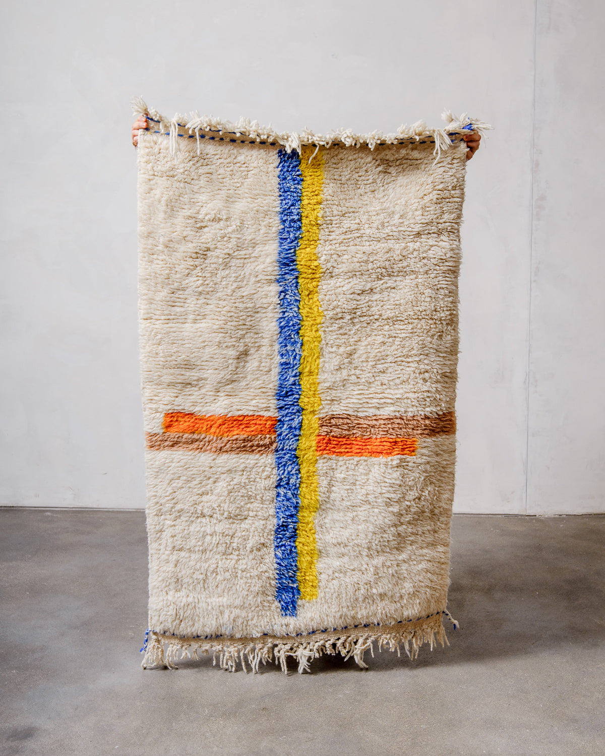 Small Berberlin Beniourain rug made of cream wool with colourful lines, being held up in front of a white wall. 