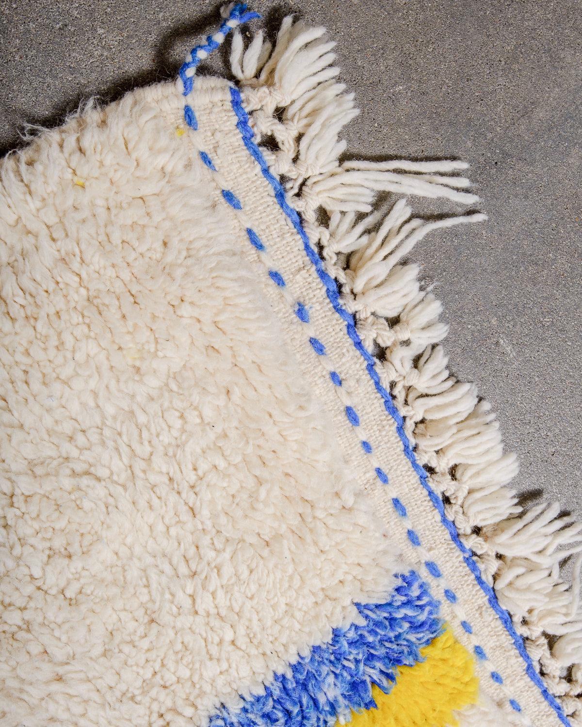 Corner of cream berber rug that is finished with tightly woven ends with blue details and natural wool tassels across the ends.