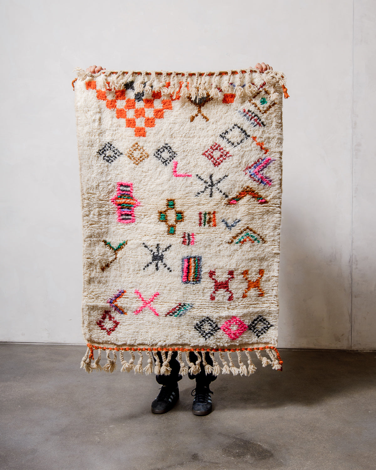 Small Berberlin Beniourain rug made of cream wool with colourful, abstract design, being held up in front of a white wall. 