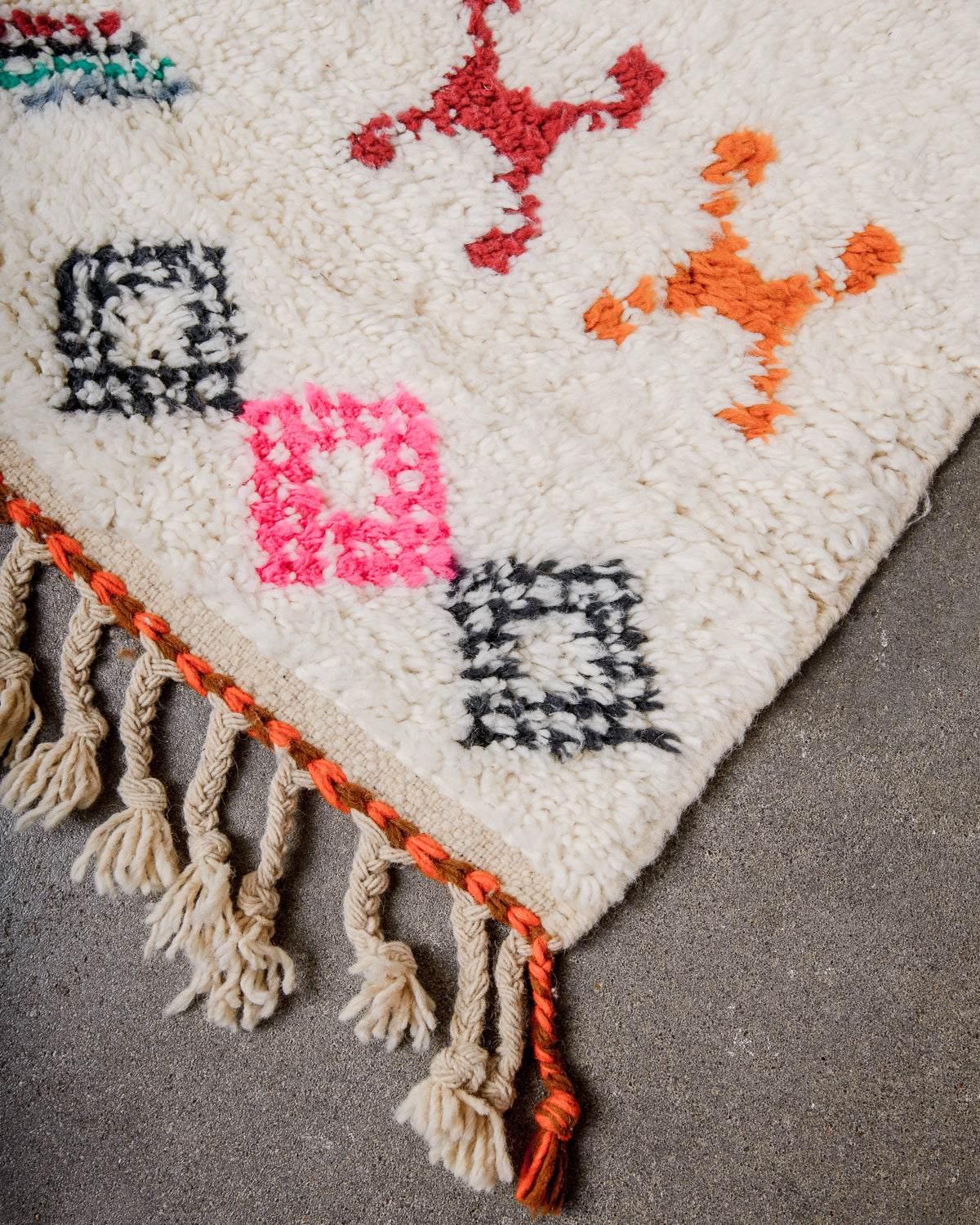 Corner of cream berber rug that is finished with tightly woven ends with orange and brown detail.