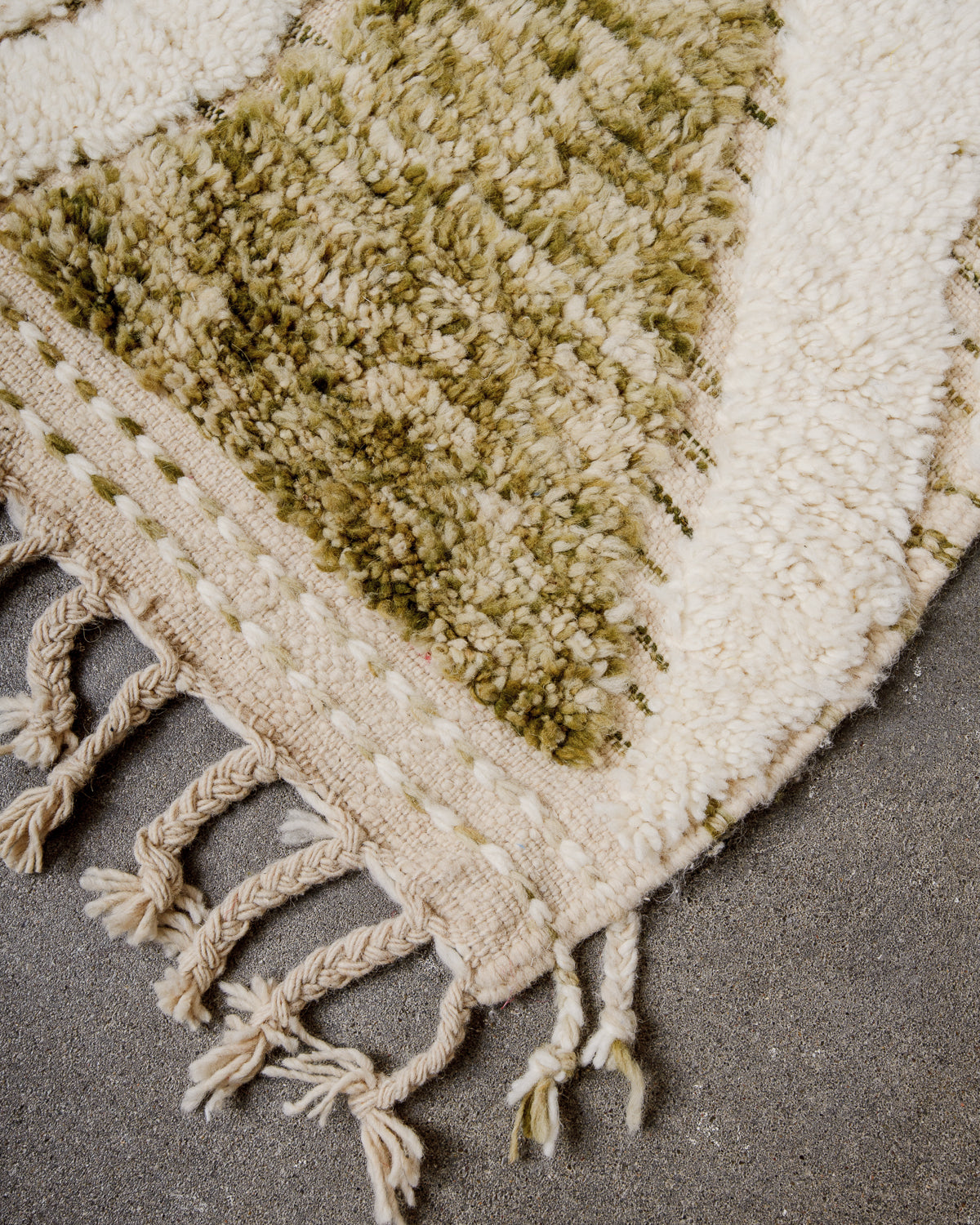 Corner of cream Beniourain wool rug with a tightly woven edge and natural wool tassels laying on concrete floor. 