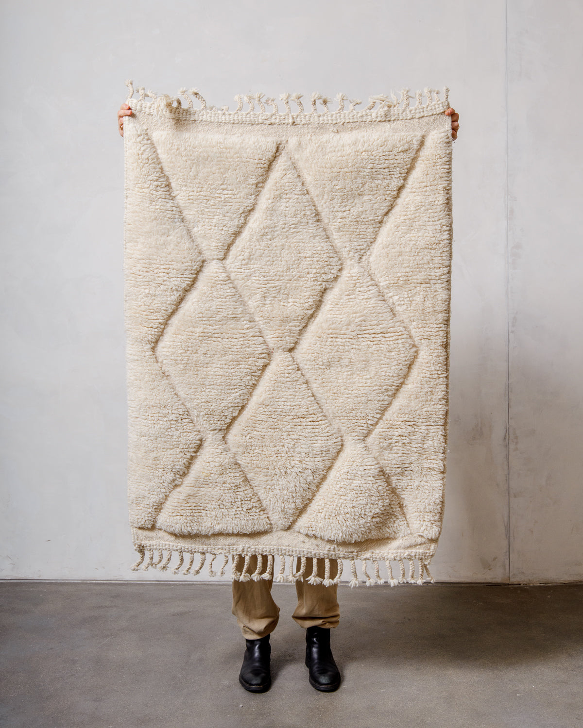 Small Berberlin Beniourain rug in cream wool with diamond design, being held up in front of a white wall. 