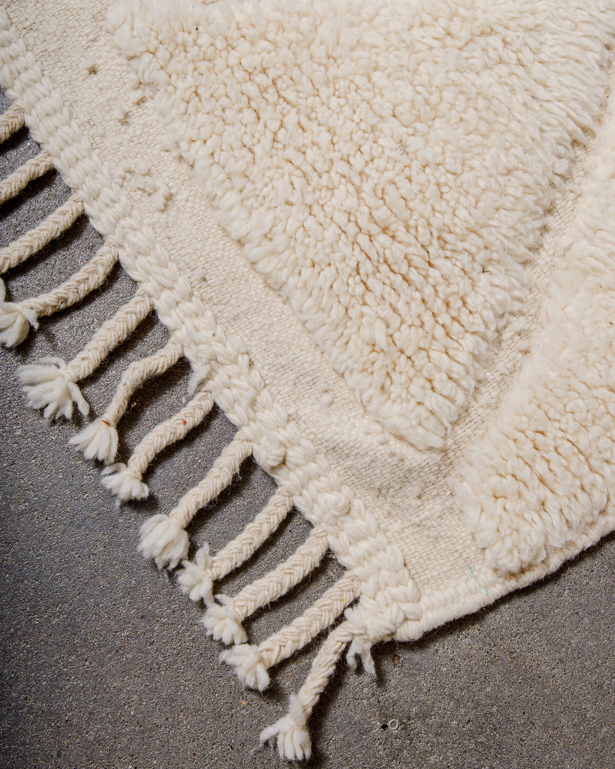 Corner of cream Beniourain wool rug with a tightly woven edge and natural wool tassels laying on concrete floor. 