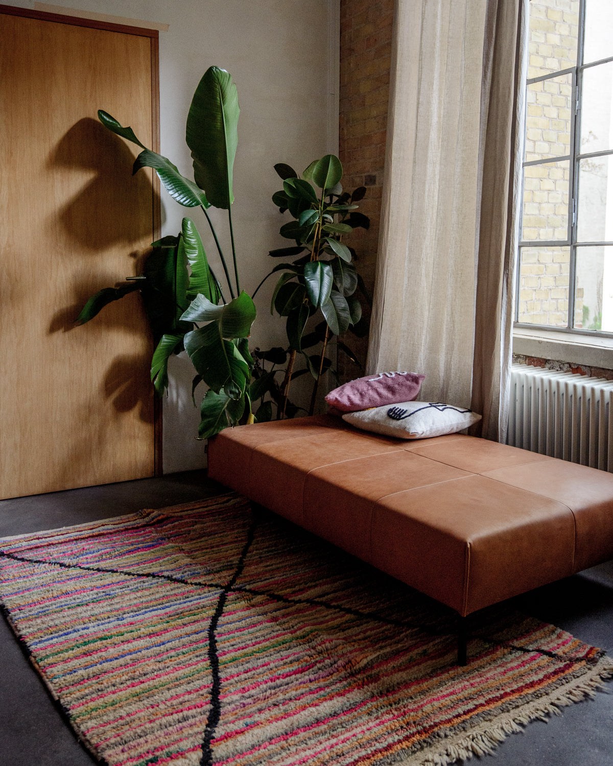 Vintage wool Berber rug on floor in front of leather Chaise Longue, indoor plants and warehouse windows.