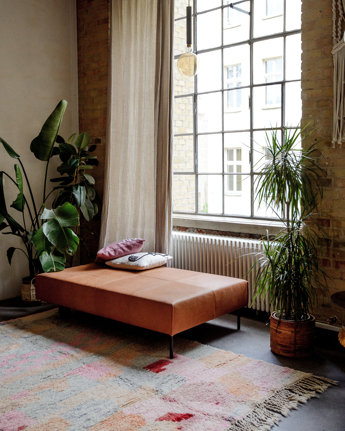 Vintage wool Berber rug on floor in front of leather Chaise Longue, indoor plants and warehouse windows.