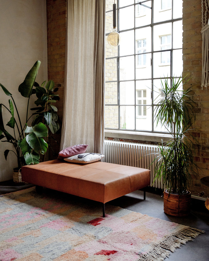 Vintage wool Berber rug on floor in front of leather Chaise Longue, indoor plants and warehouse windows.