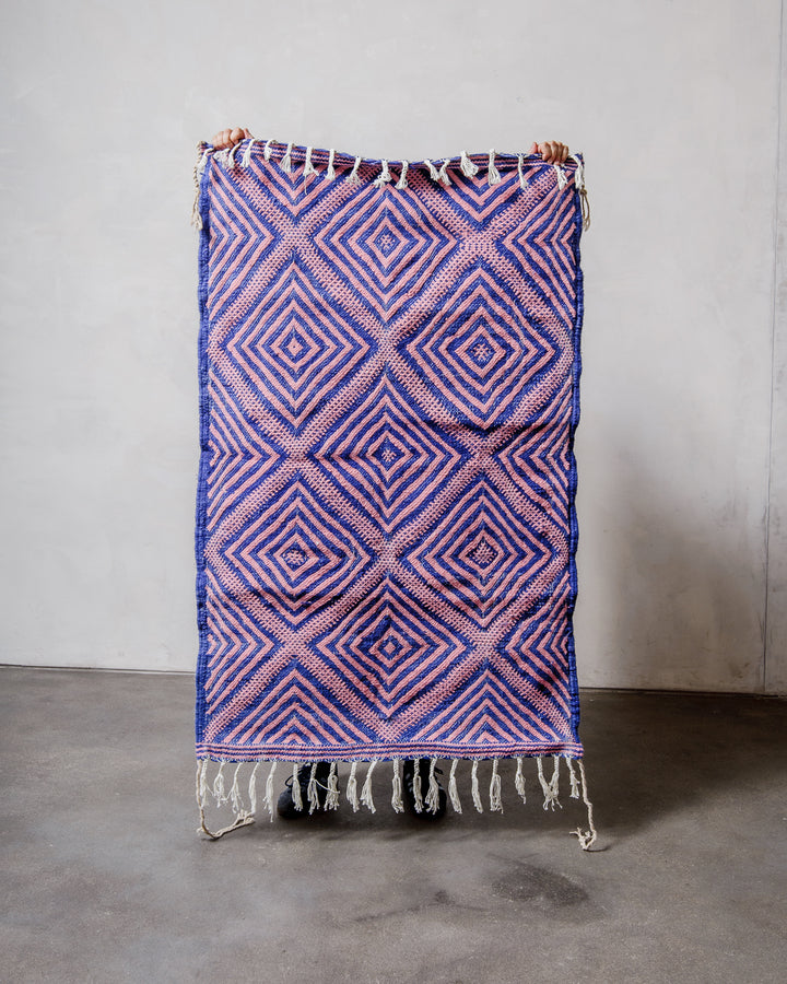 Small Berberlin Kelim Rug in pink and blue being held up in front of a white wall.