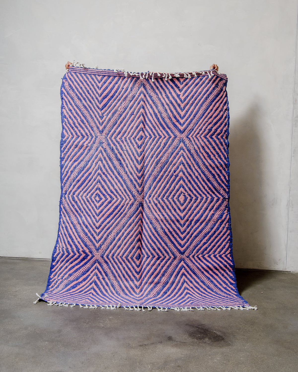 Berberlin Kelim rug made of blue and pink wool being held up in front of a white wall. 