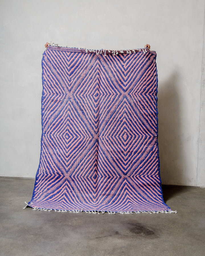 Berberlin Kelim rug made of blue and pink wool being held up in front of a white wall. 
