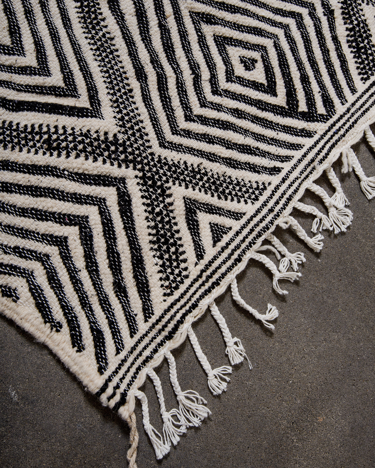 Corner of Kelim Rug in black and white with white cotton tassels, laying on concrete.