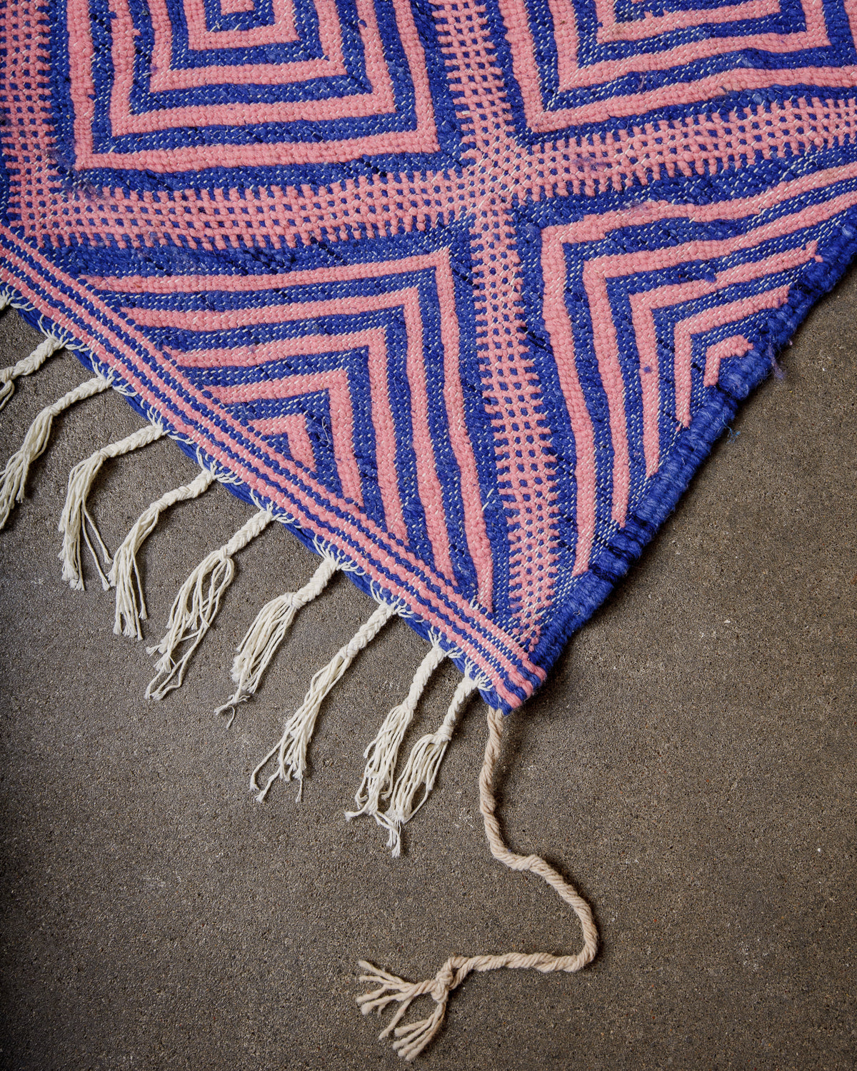 Corner of Kelim Rug in pink and blue with white cotton tassels, laying on concrete.