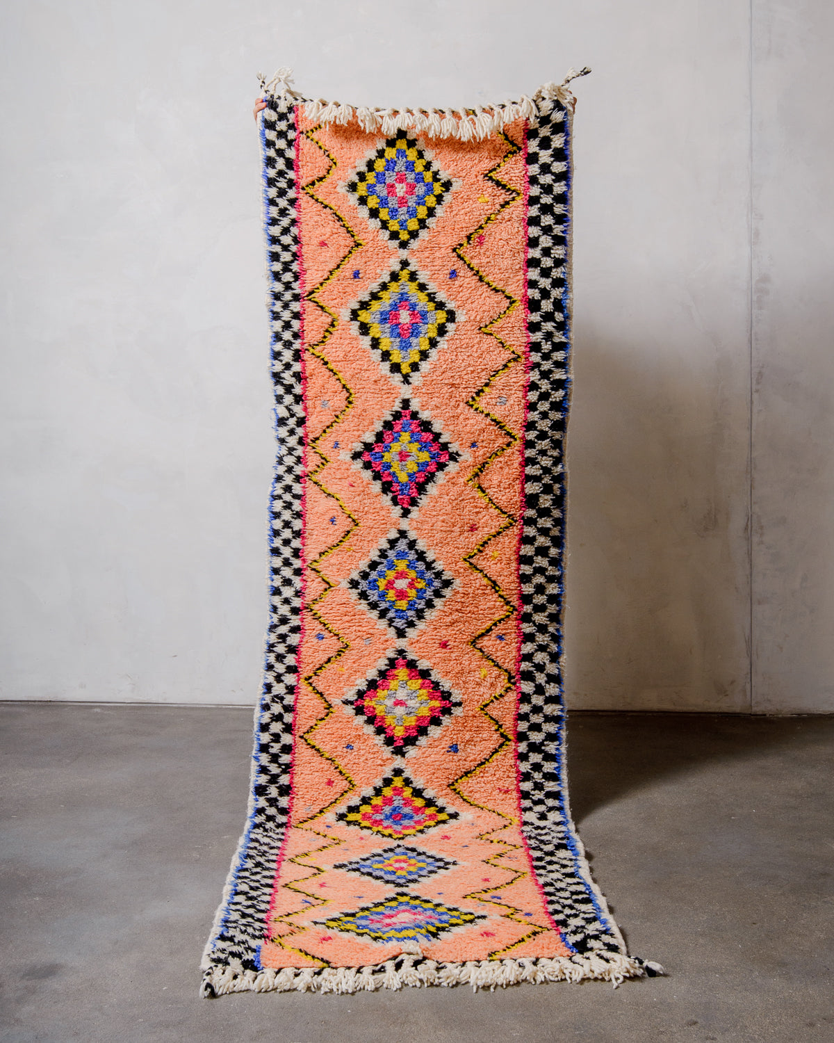Berberlin runner rug made of sheep's wool, being held up in front of a white wall and concrete floor. 