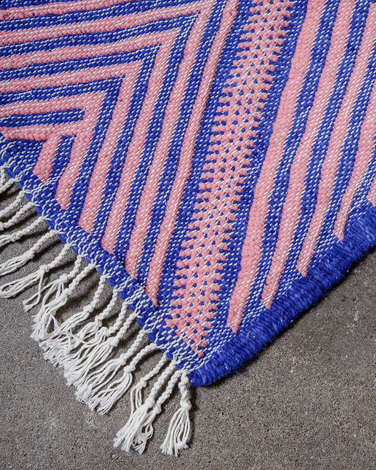 Corner of Kelim Rug in pink and blue with white cotton tassels, laying on concrete.