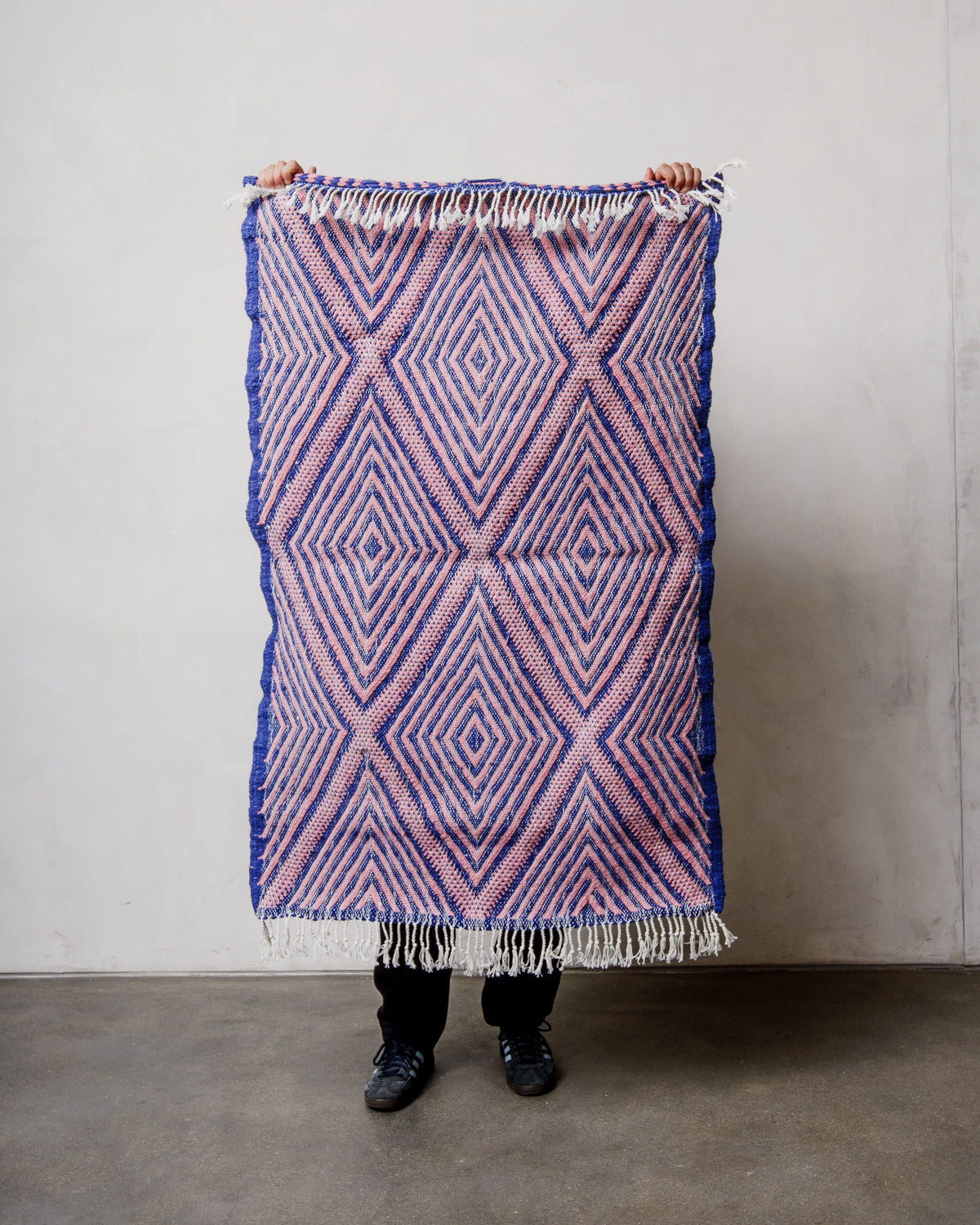 Small Berberlin Kelim Rug in pink and blue being held up in front of a white wall.