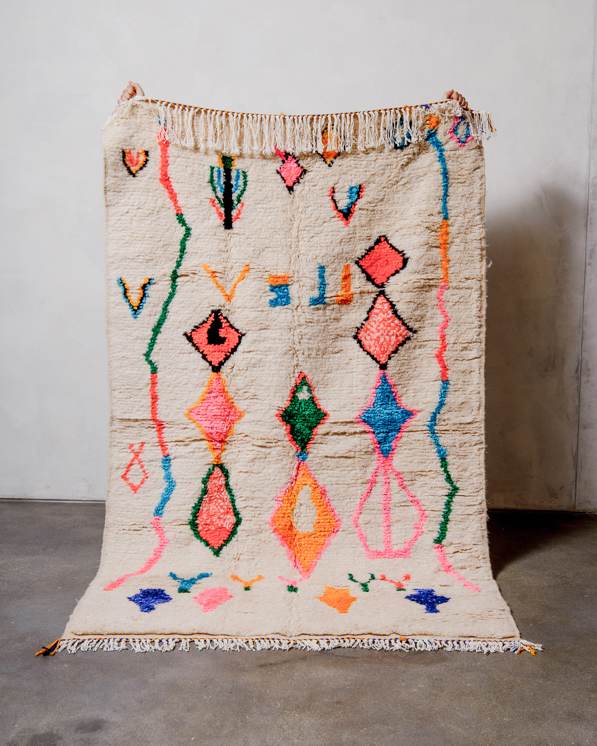 Berberlin Azilal rug made of fluffy cream wool and colourful design, held up  in front of a white wall and cement floor.
