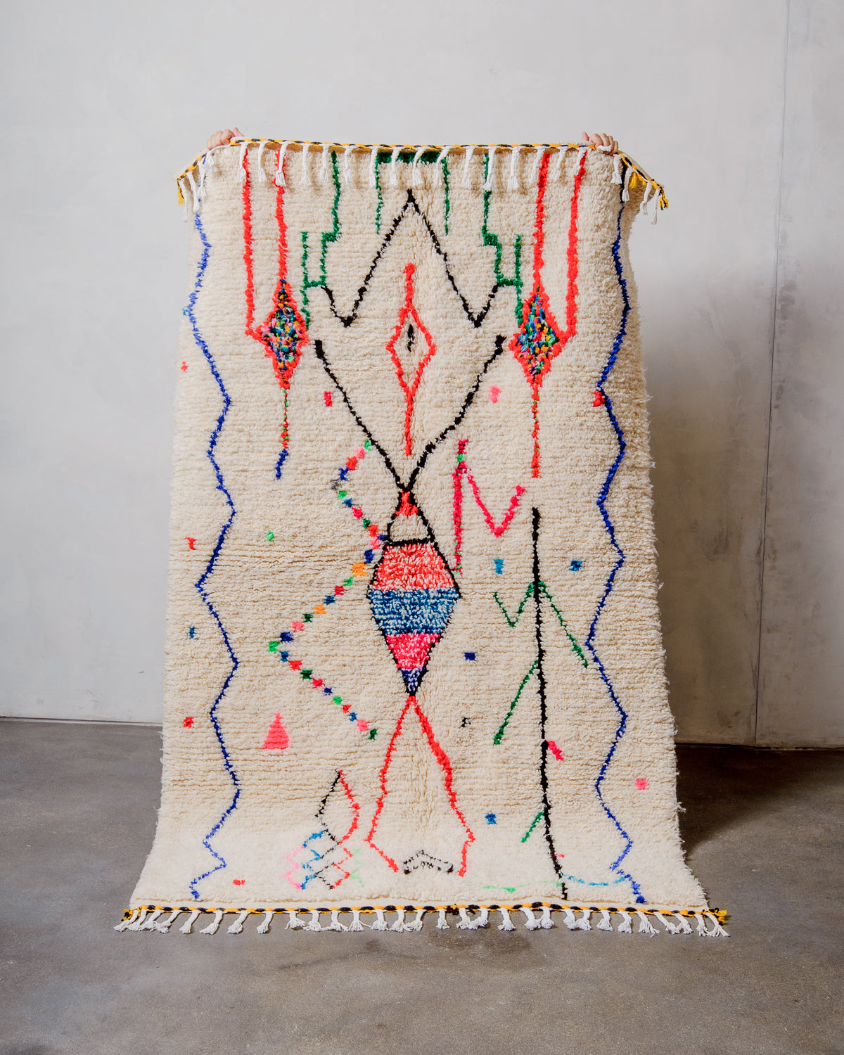 Berberlin Azilal rug made of fluffy cream wool and colourful design, held up in front of a white wall and cement floor.