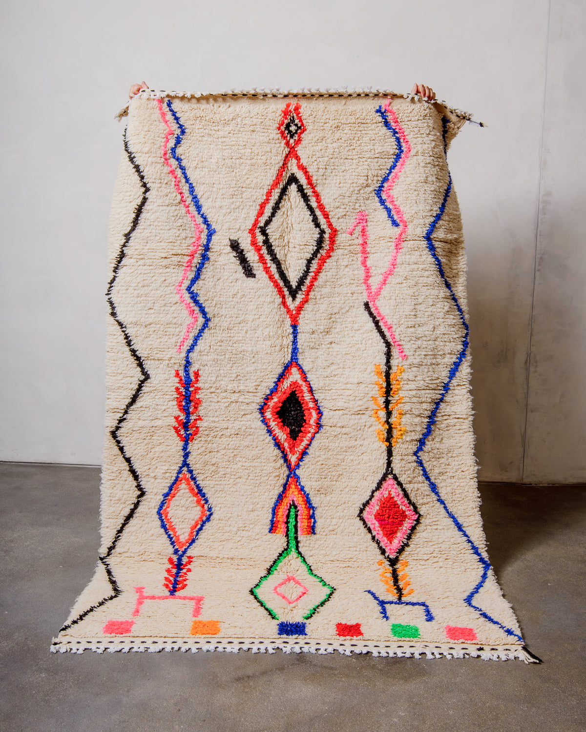 Berberlin Azilal rug made of fluffy cream wool and colourful design, held up  in front of a white wall and cement floor.