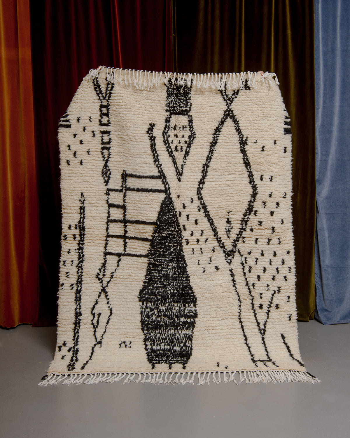 Azilal rug with abstract design in black being held up in front of a wall of velvet curtains.