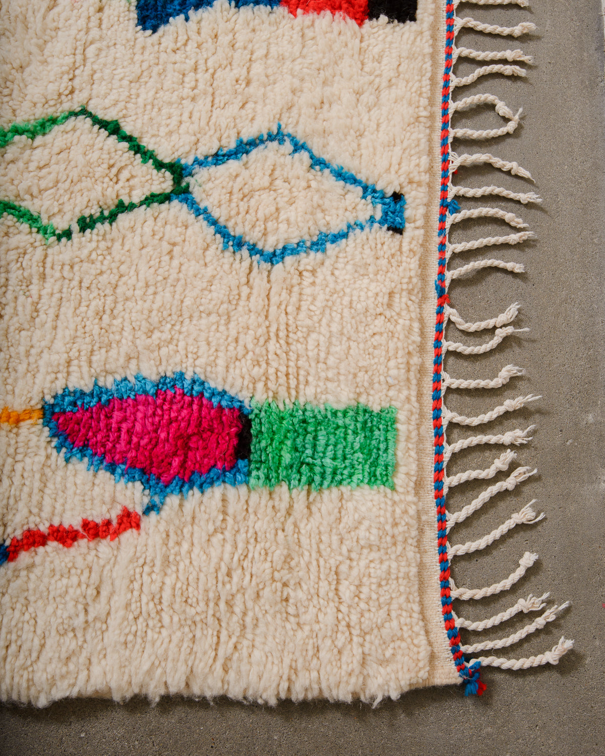Corner of Azilal rug with cream wool and white cotton tassels and blue and pink weaving across the edge.
