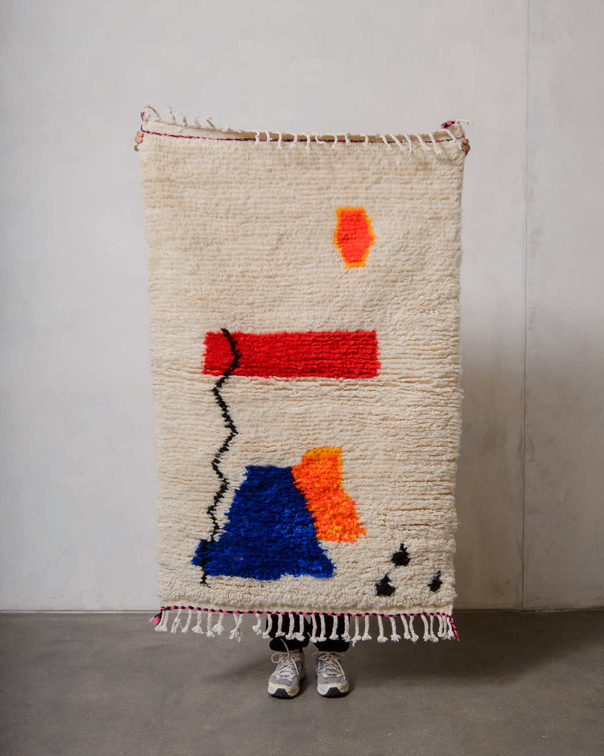 Berberlin Azilal rug in cream wool with colourful cotton  being held up in front of a white wall.