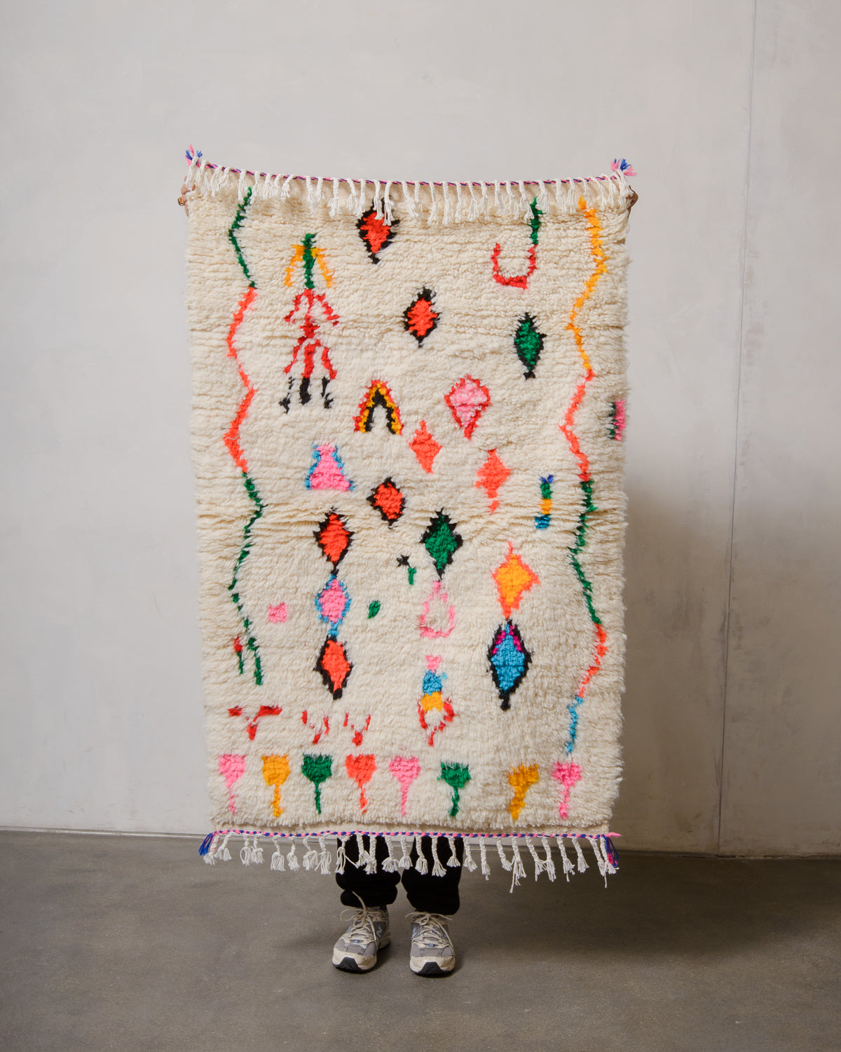 Berberlin Azilal rug in cream wool with colourful cotton  being held up in front of a white wall.