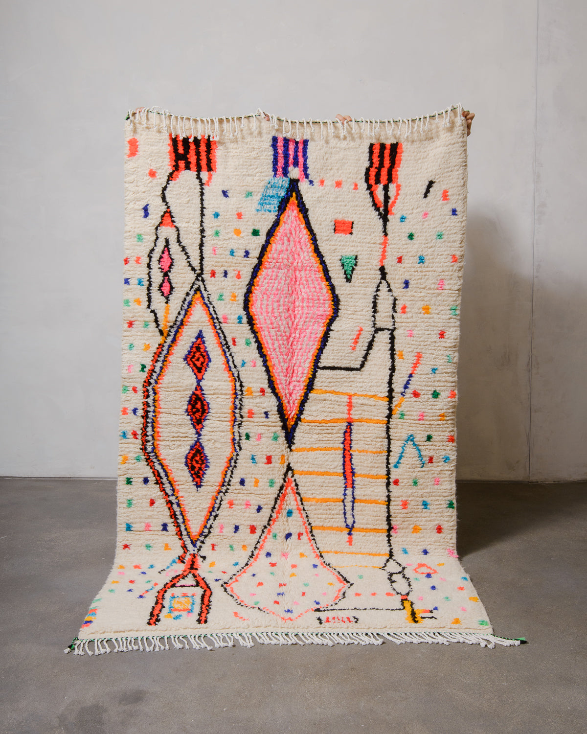 Berberlin Azilal rug made of fluffy cream wool and colourful design, held up in front of a white wall and cement floor.