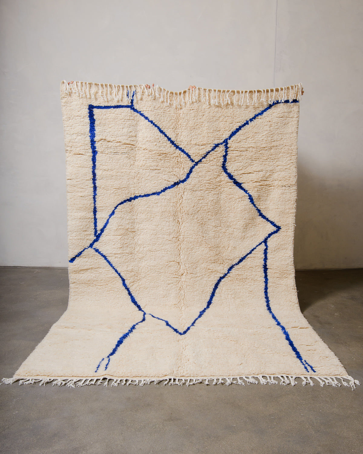 Berberlin Azilal rug made of fluffy cream wool and a minimalist blue line design, held up in front of a white wall and cement floor.