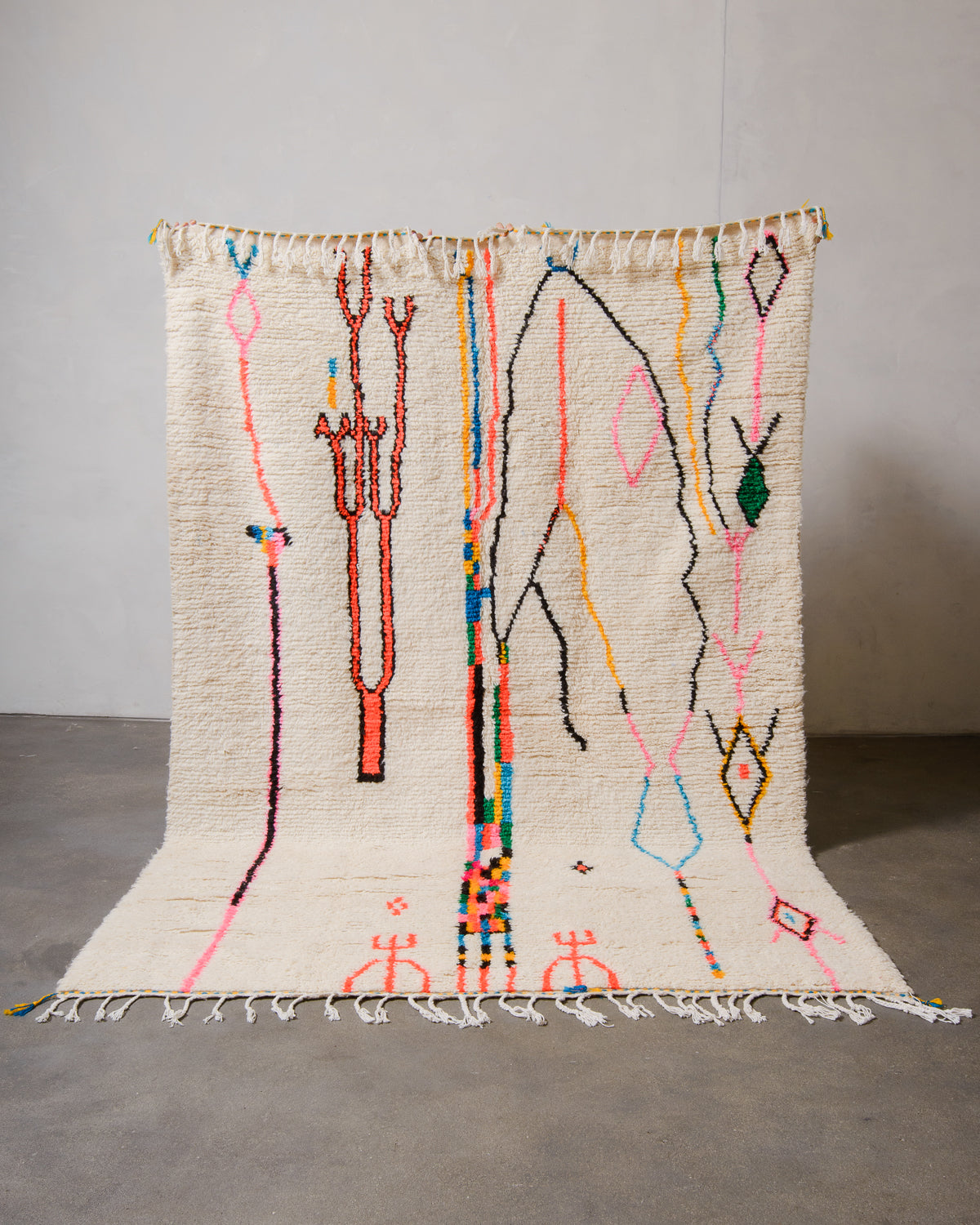 Berberlin Azilal rug made of fluffy cream wool and colourful design, held up in front of a white wall and cement floor.