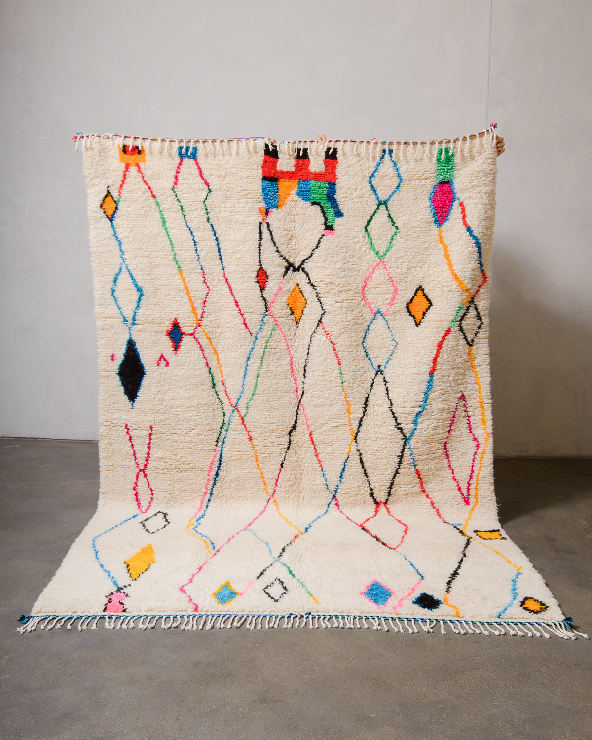 Berberlin Azilal rug made of fluffy cream wool and colourful design, held up in front of a white wall and cement floor.