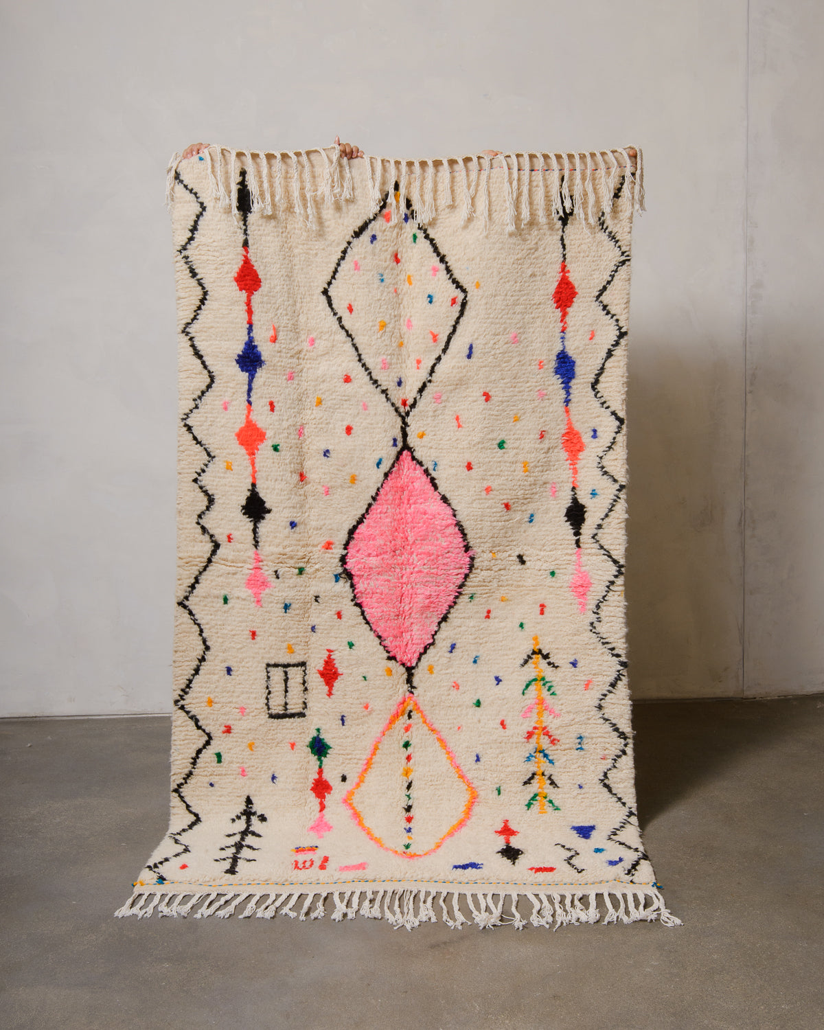 Berberlin Azilal rug made of fluffy cream wool and colourful design, held up in front of a white wall and cement floor.
