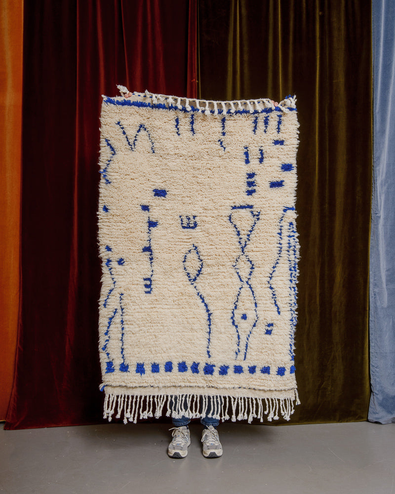 Berberlin Azilal rug with blue motifs held in front of a velvet curtain.
