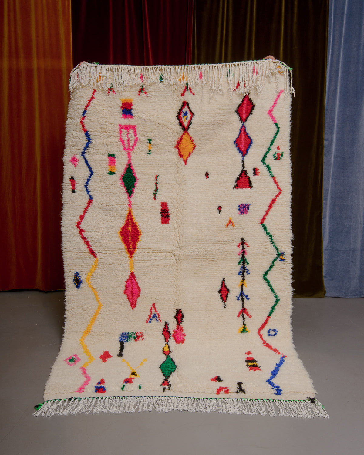 Berberlin azilal rug with colourful design being held in front of velvet curtains.