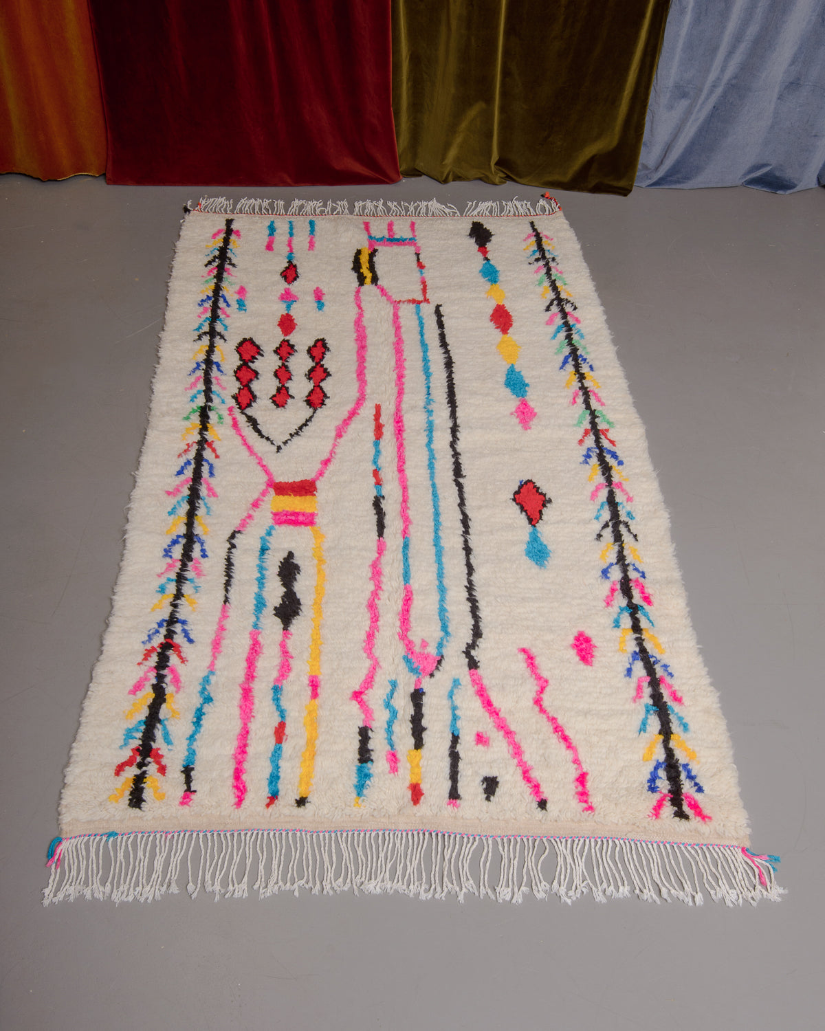 Azilal rug with colourful, abstract design laid flat on grey floor in front of a wall of velvet curtains.