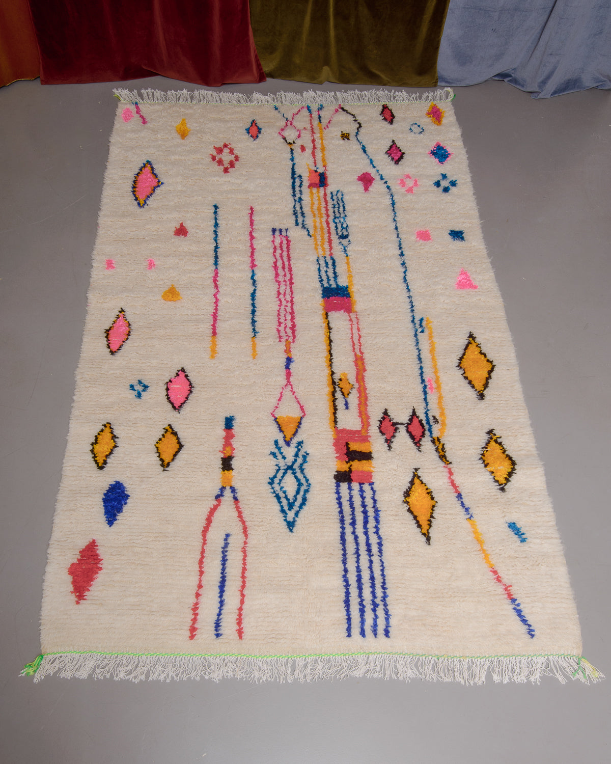 Azilal rug with colourful, abstract design laid flat on grey floor in front of a wall of velvet curtains.