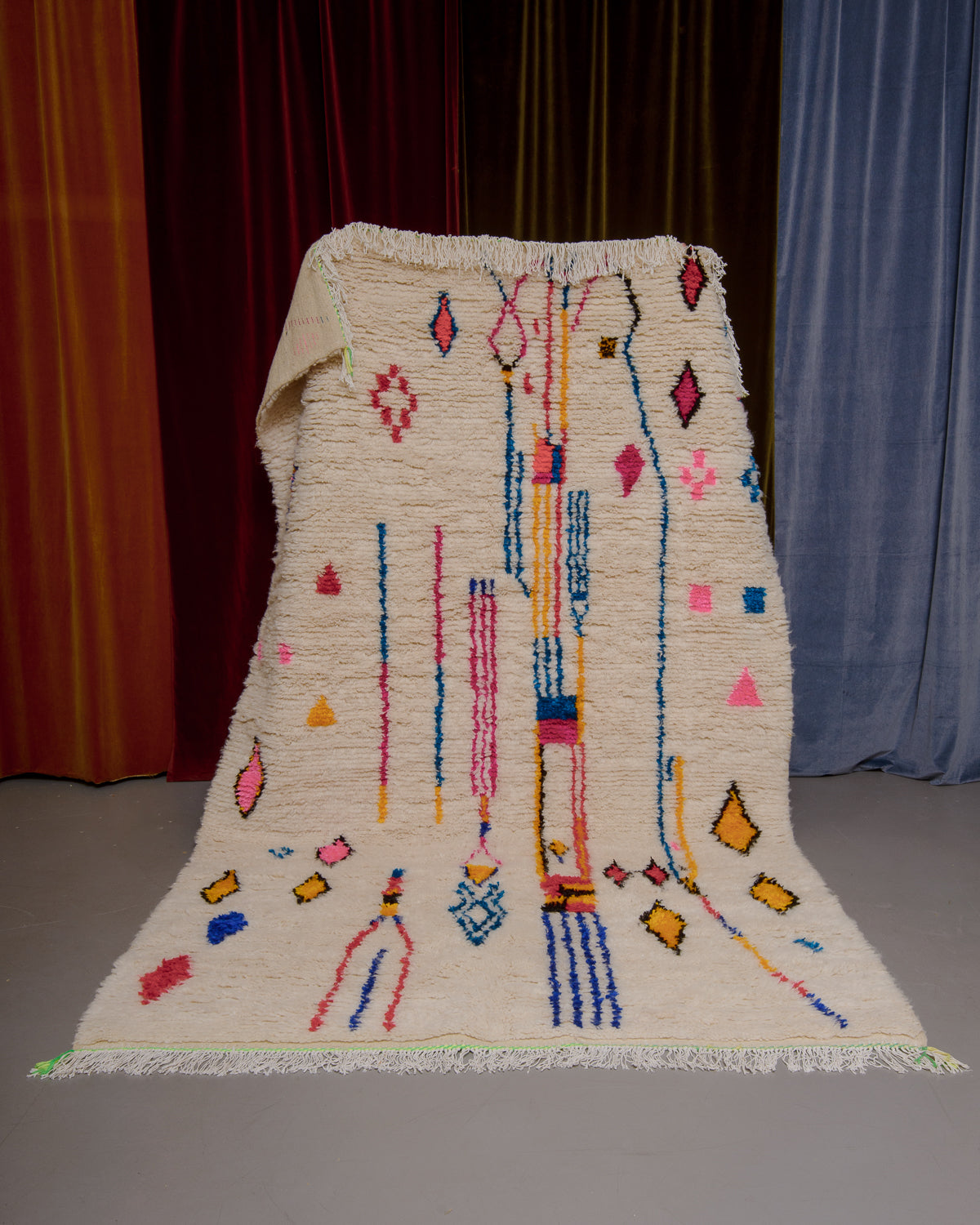 Azilal rug with colourful, abstract design being held up in front of a wall of velvet curtains.