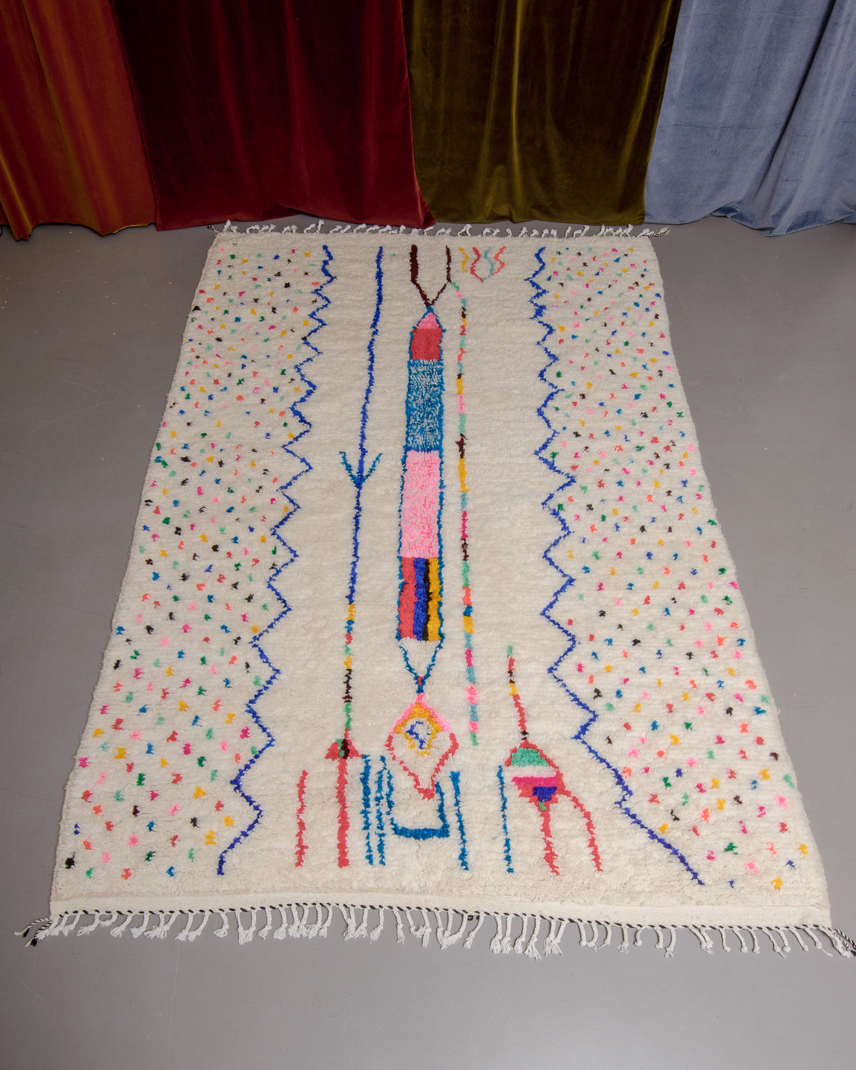 Azilal rug with colourful, abstract design laid flat on grey floor in front of a wall of velvet curtains.
