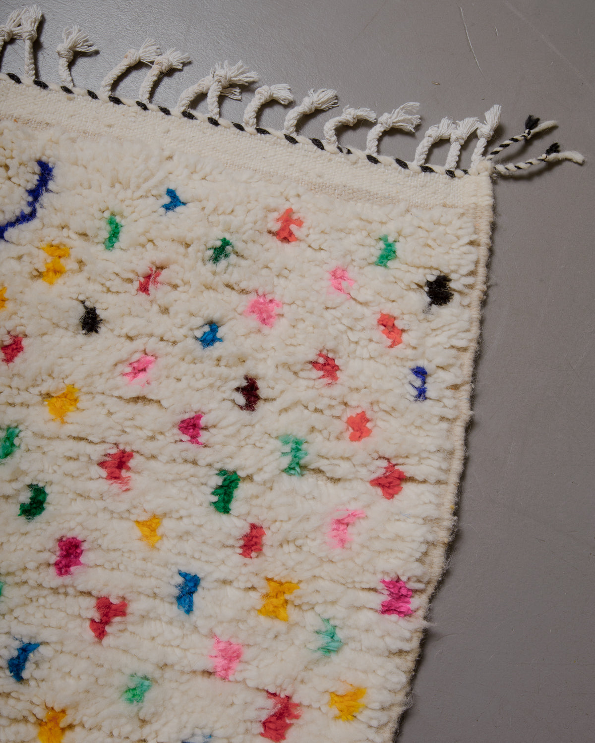 Corner of azilal rug with white cotton tassels, colourful polka dots and black and white stitching across the end. 
