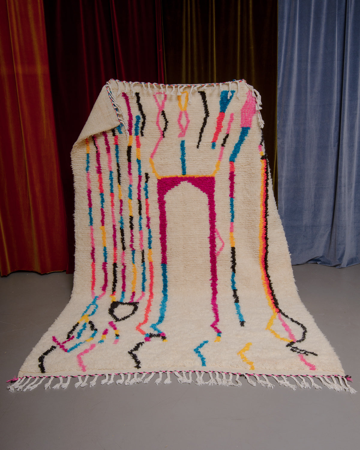 Azilal rug with colourful, abstract design being held up in front of a wall of velvet curtains.