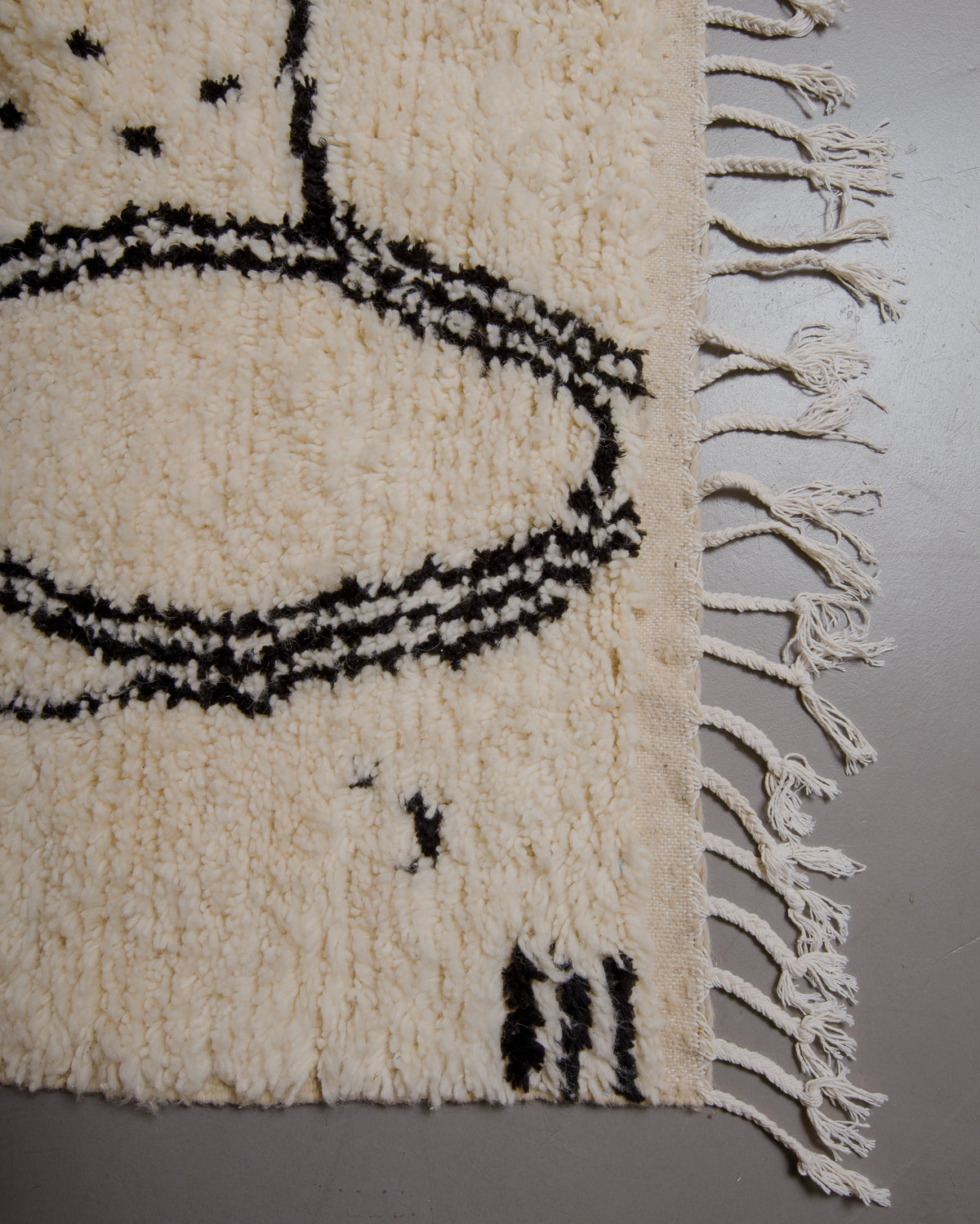 Corner of azilal rug with white cotton tassels on grey floor. 