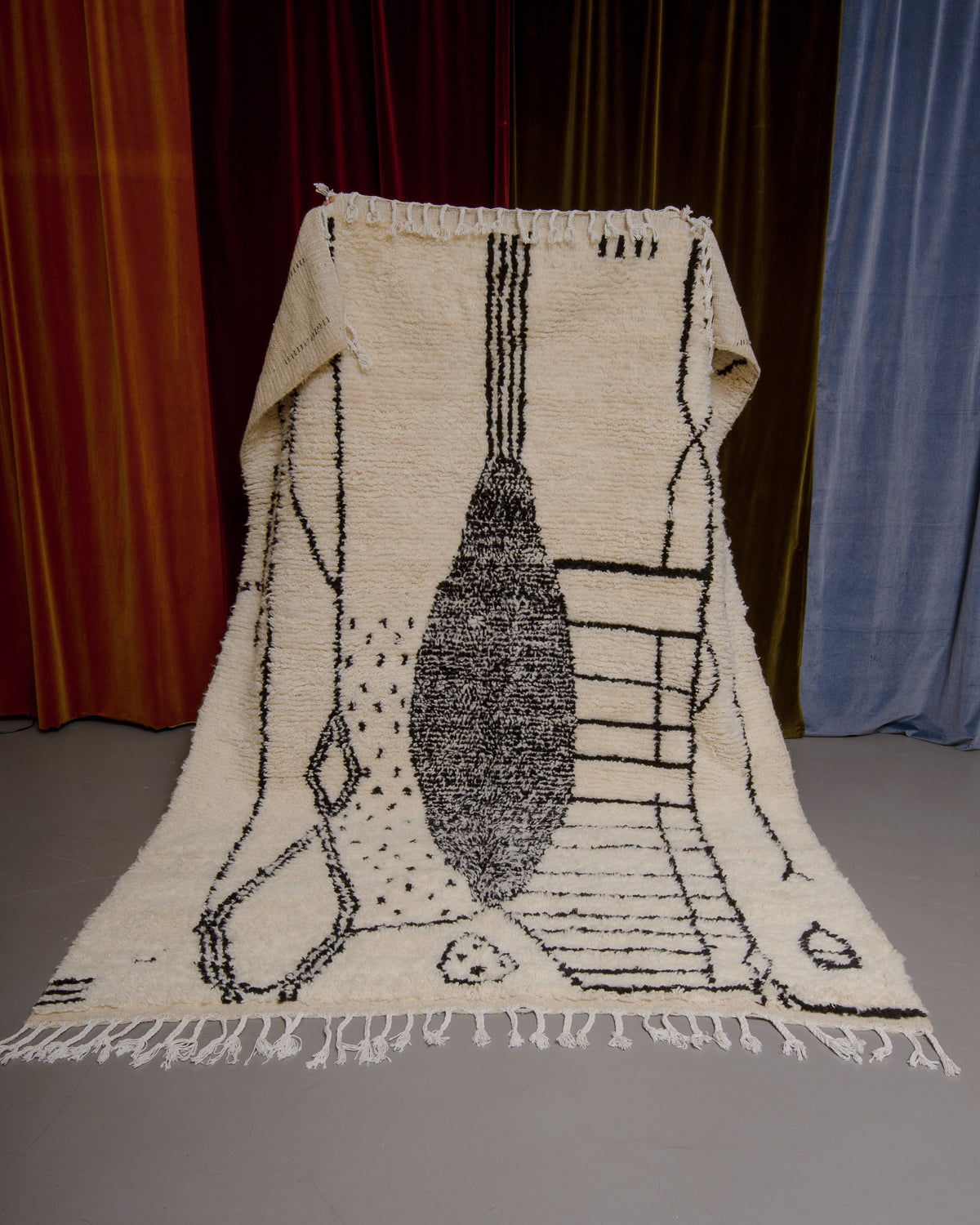 Azilal rug with abstract design in black being held up in front of a wall of velvet curtains.