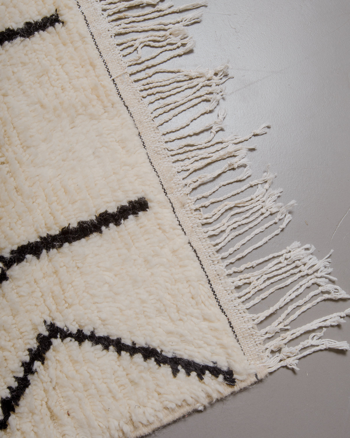 Corner of azilal rug with white cotton tassels on grey floor. 