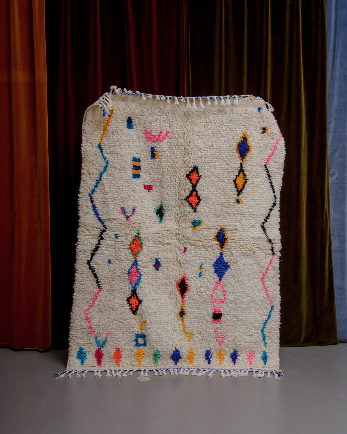 Azilal rug with colourful, abstract design being held up in front of a wall of velvet curtains.