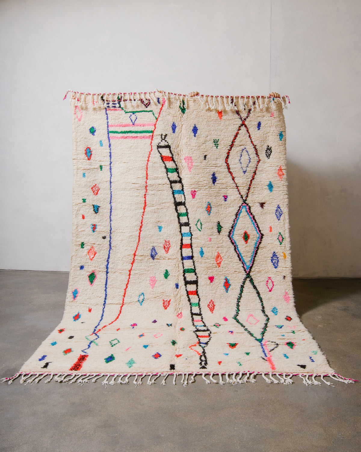 Berberlin Azilal rug made of fluffy cream wool and colourful design, held up in front of a white wall and cement floor.
