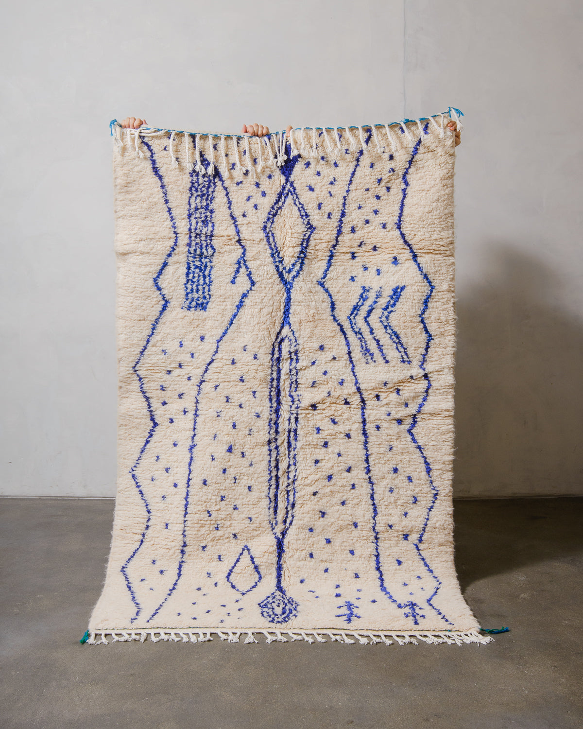 Berberlin Azilal rug made of fluffy cream wool and abstract design in cobalt blue, held up in front of a white wall and cement floor.