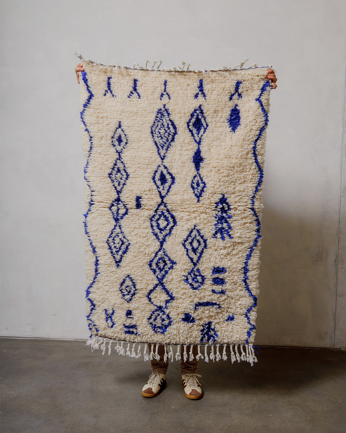Person holding a blue and cream patterned rug against a plain wall.