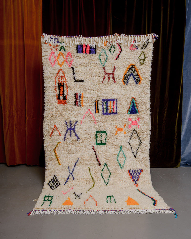 Berberlin Azilal rug with cream wool and colourful symbols being held up in front of velvet curtains.